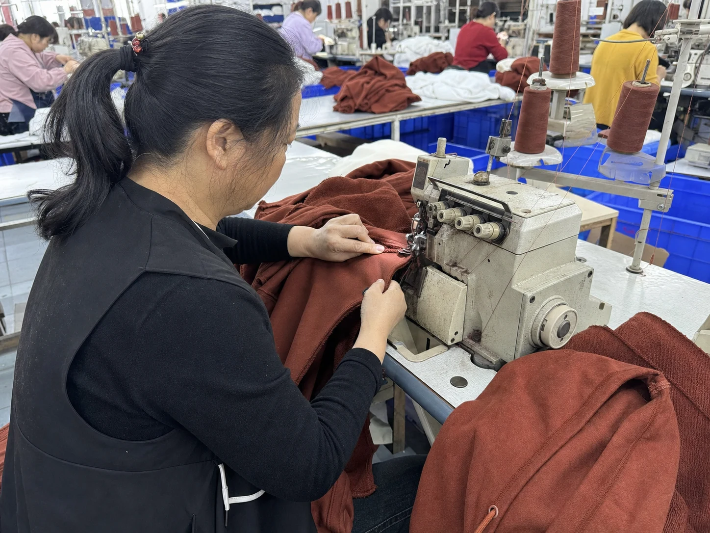 An operator joins the sleeve and body of a rust-coloured hoodie at an industrial sewing station.