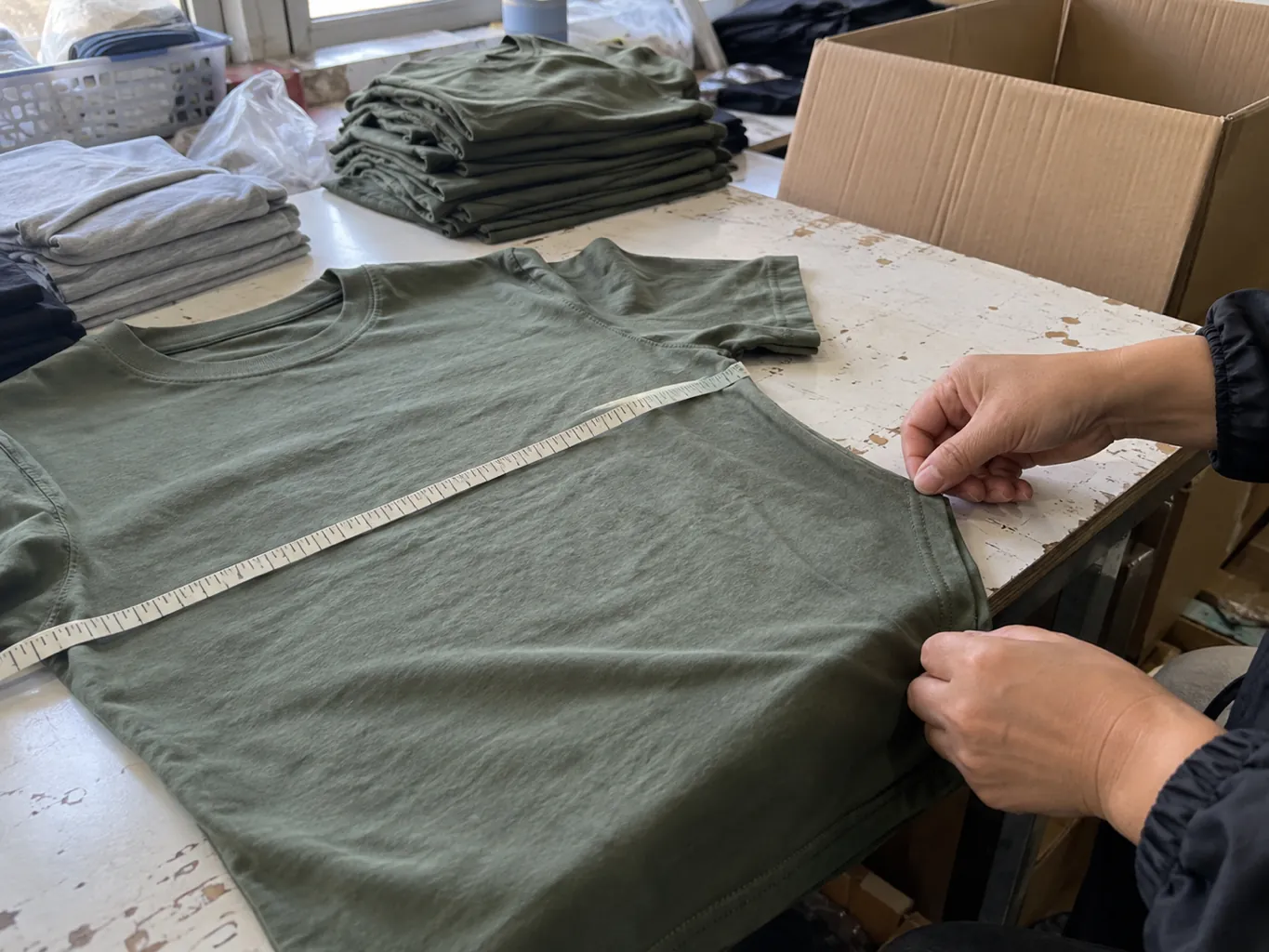 Mains vérifiant une couture de chemise à côté d’un mètre ruban et d’un carton ouvert.