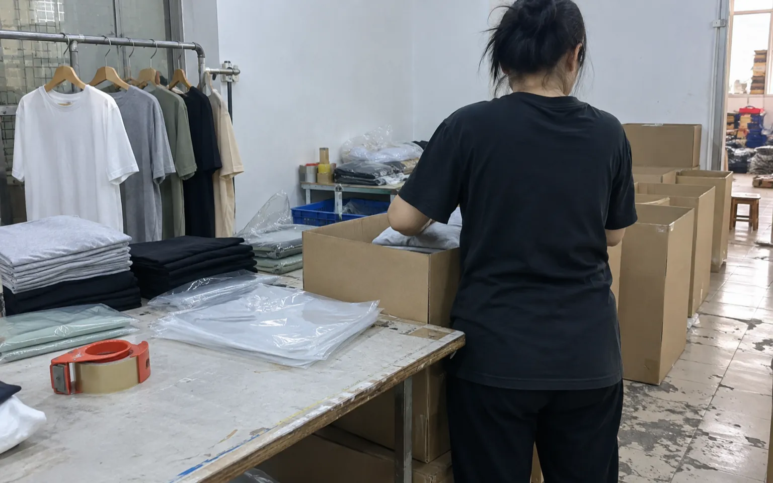 Ouvrier emballant des vêtements pliés dans des cartons d’expédition sans marquage, à côté d’échantillons de vêtements suspendus.