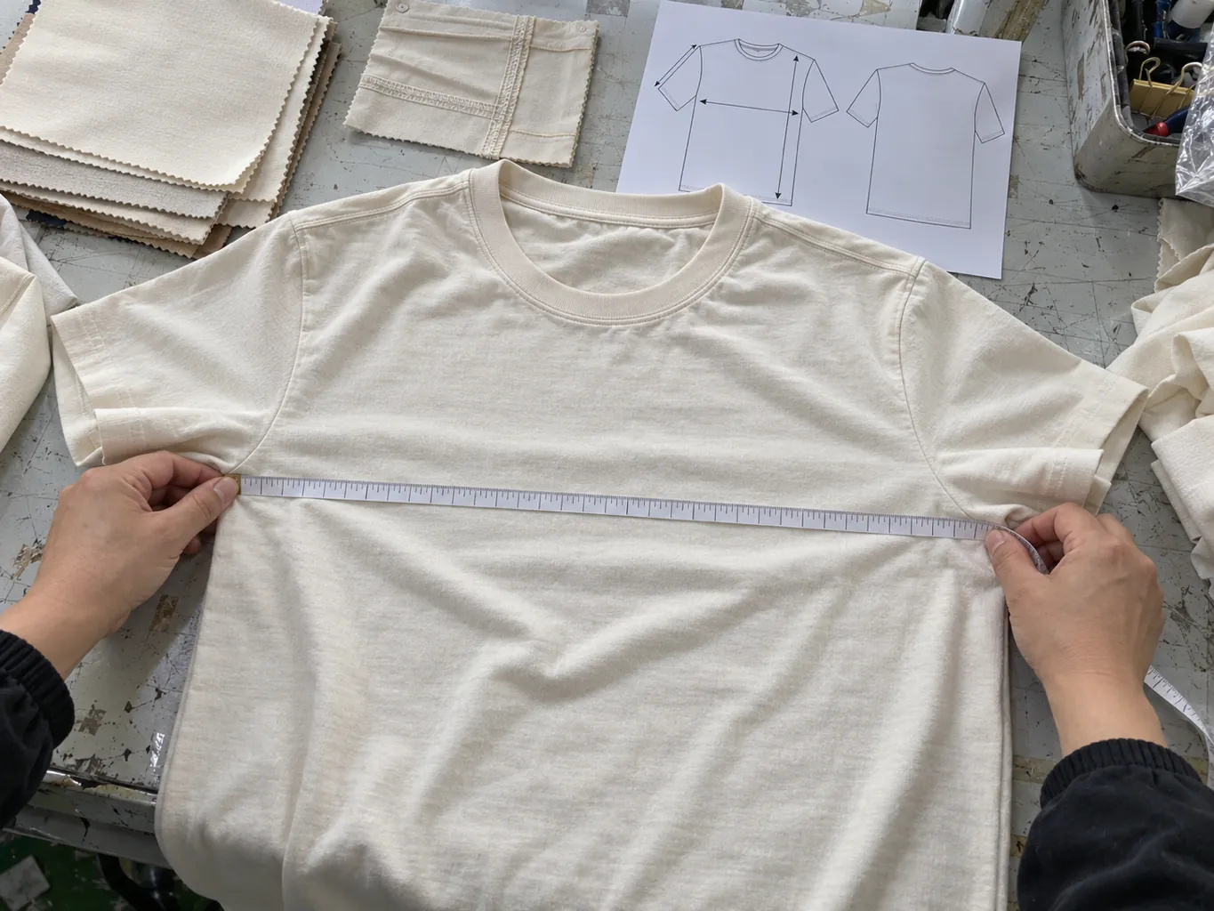 Hände messen die Brustweite eines Shirts neben Stoffmustern und einer technischen Zeichnung.