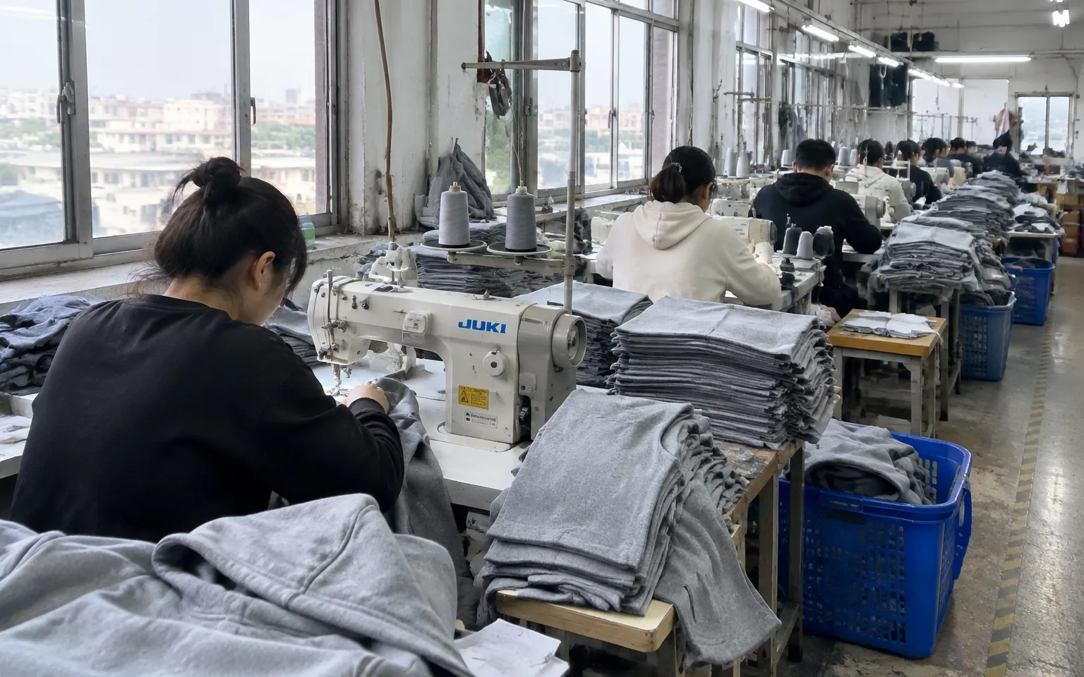 Hoodie production line in a Guangzhou garment factory, with stacks of cut grey fleece panels beside the sewing machines